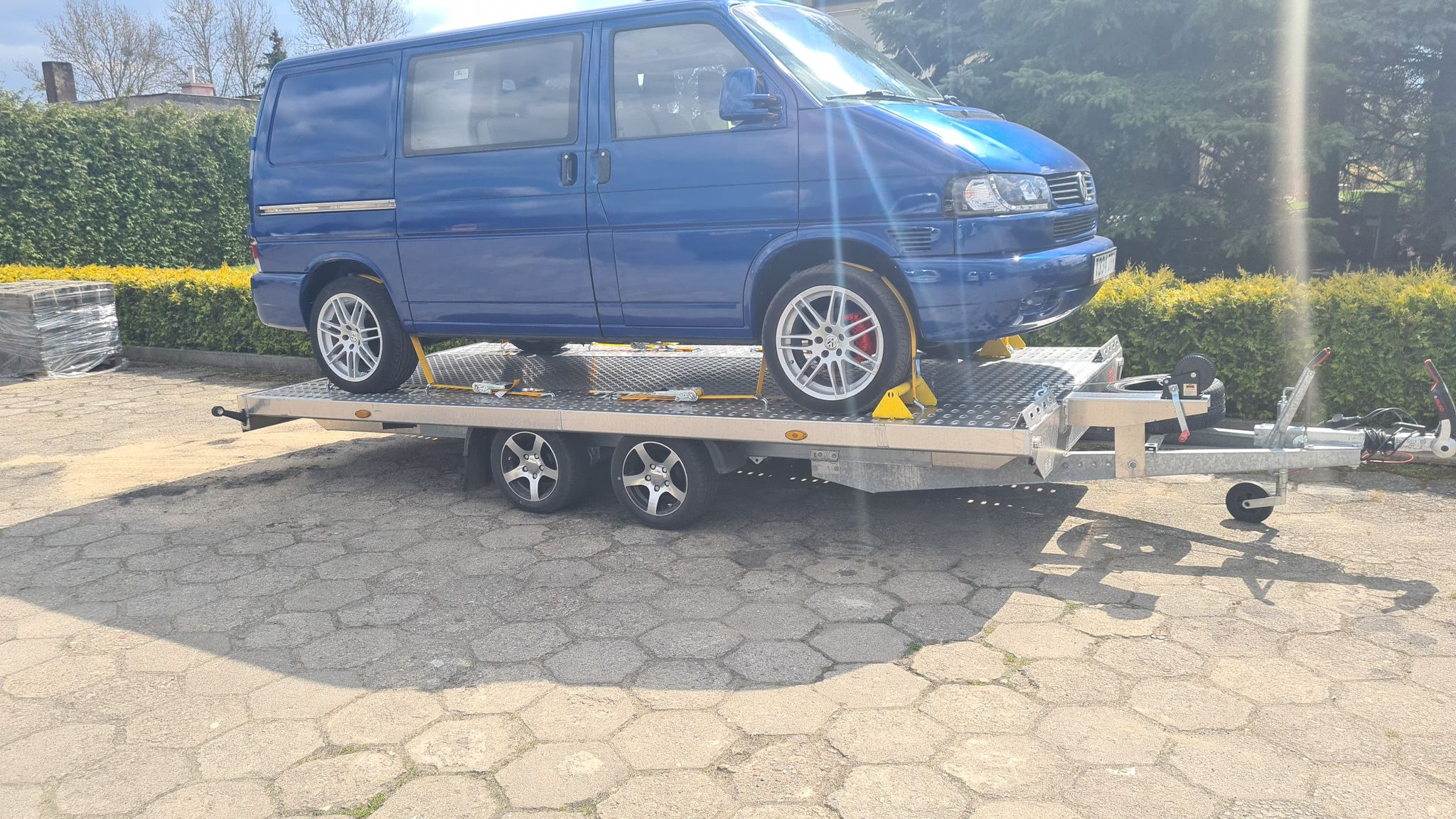 Niebieski van Volkswagen T4 na srebrnej, dwuosiowej lawecie z żółtymi klinami pod kołami, widok z boku na tle zielonych krzewów i kostki brukowej.