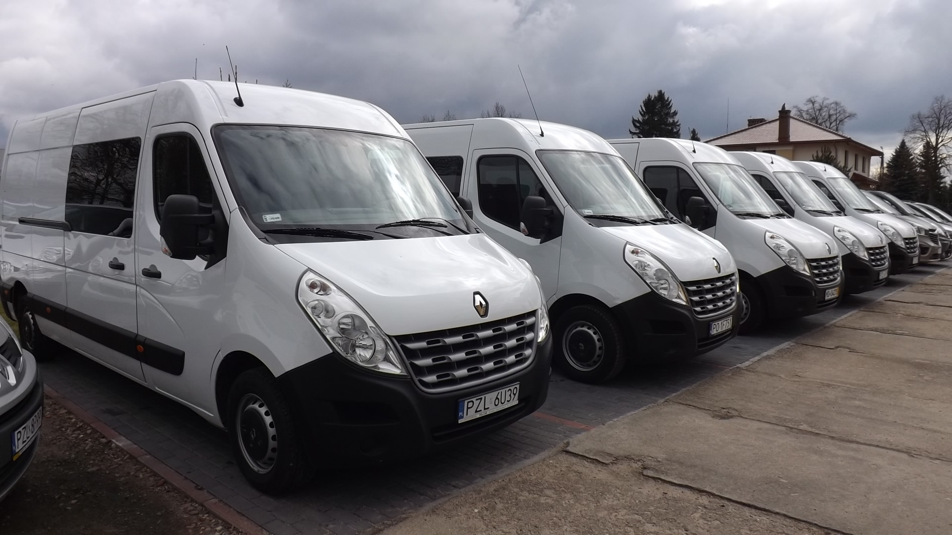 Rząd białych samochodów dostawczych Renault Master zaparkowanych na betonowym placu, widoczne numery rejestracyjne, pochmurne niebo w tle.