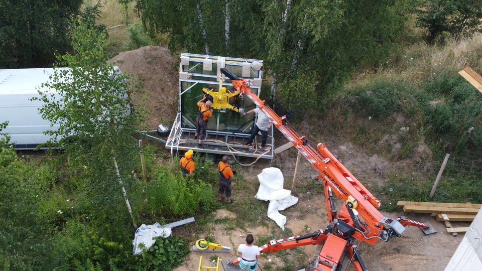 Montaż okna panoramicznego za pomocą dźwigu Minizurawie Ogrodowski, widok z góry na ekipę montażową w strojach roboczych i kaskach, biały bus w tle.