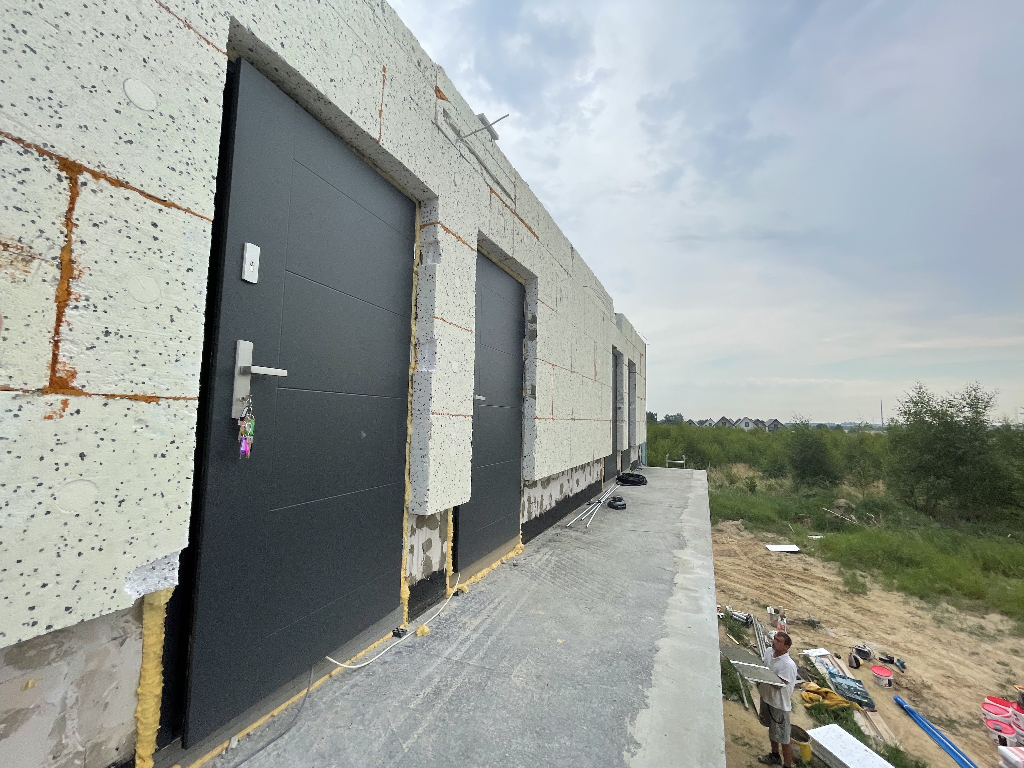 Szara brama garażowa z kluczami w zamku, zamontowana w budynku w trakcie ocieplania elewacji styropianem, widoczne niedokończone prace i narzędzia.