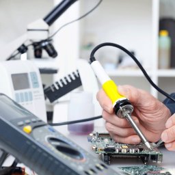 DEADLINK P.S.A. - Elektronik lutuje płytkę drukowaną przy użyciu lutownicy z żółtą rączką, obok multimetr, oscyloskop i mikroskop.