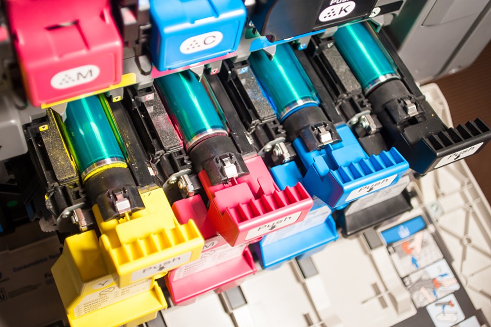 Wymiana tonerów CMYK w drukarce laserowej, widoczne wkłady z oznaczeniami kolorów i instrukcja obsługi na obudowie.