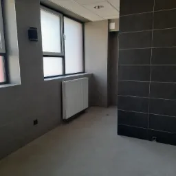 Wnętrze łazienki po remoncie z szarymi płytkami na ścianach i podłodze, biały grzejnik pod oknem z matową szybą, widoczny fragment kabiny prysznicowej.