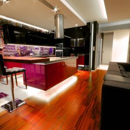 Polska Fabryka Mebli - Nowoczesna kuchnia z wyspą o bordowych frontach, drewnianym blatem i fototapetą z widokiem na miasto, oświetlona listwami LED pod szafkami i na podłodze, widoczny fragment drewnianej podłogi...