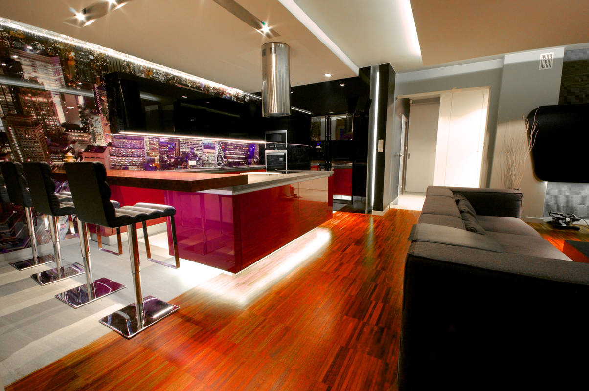 Nowoczesna kuchnia z wyspą o bordowych frontach, drewnianym blatem i fototapetą z widokiem na miasto, oświetlona listwami LED pod szafkami i na podłodze, widoczny fragment drewnianej podłogi...