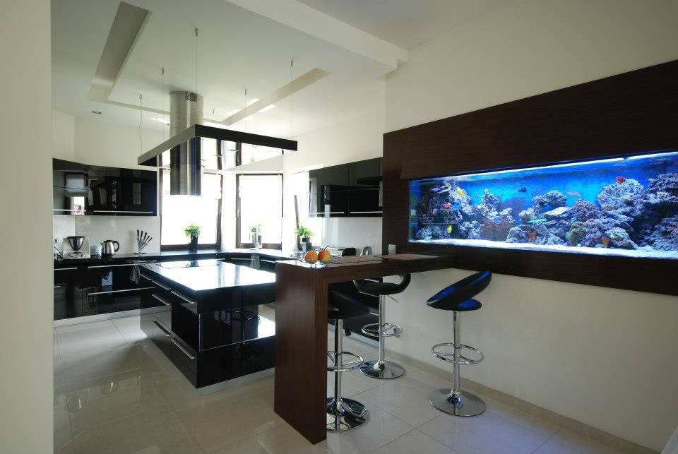 Nowoczesna kuchnia z czarnymi szafkami i wyspą, oświetlona wiszącymi lampami, zintegrowana z barkiem i dużym akwarium w zabudowie.