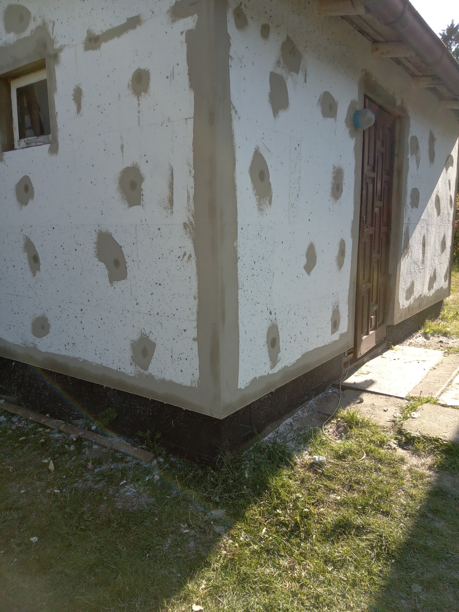 Ocieplony budynek z widocznymi łatami zaprawy, przygotowany do nałożenia tynku, z fragmentem tęczy na trawie.