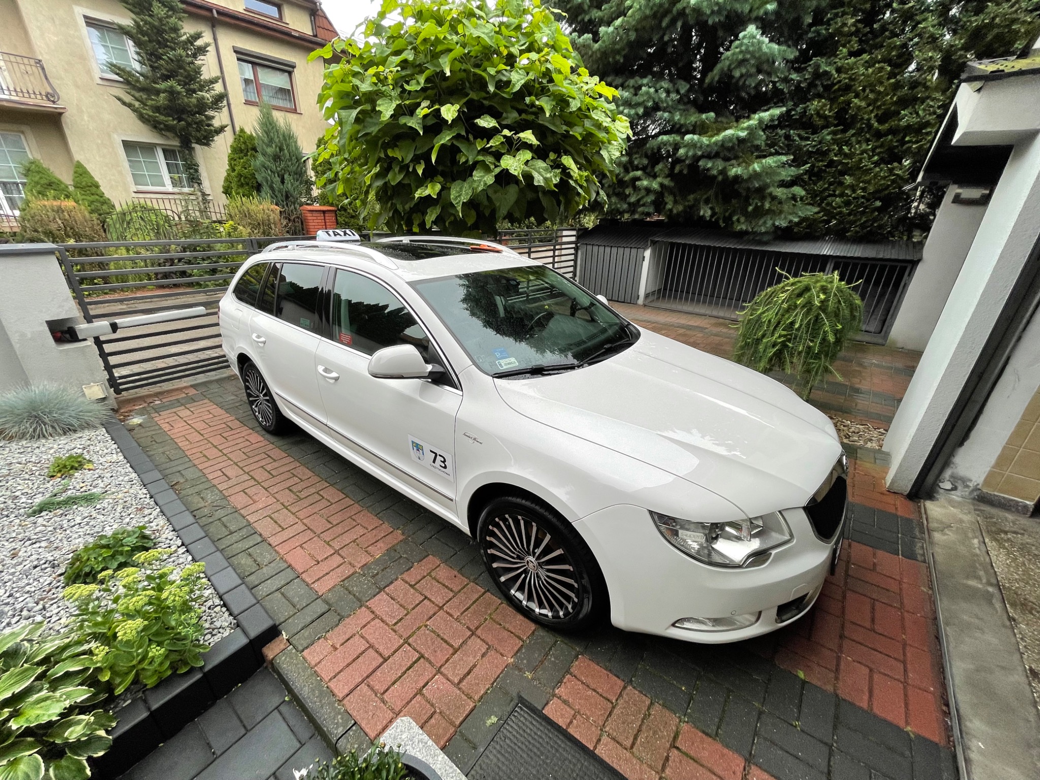 Białe kombi taxi marki Skoda Superb zaparkowane na brukowanym podjeździe przed domem z ogrodem, widoczny napis TAXI na dachu i logo firmy na drzwiach.