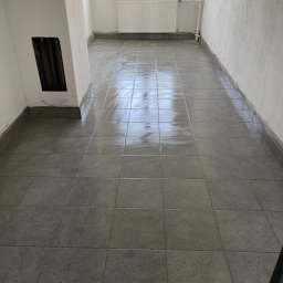 Remonty Dobry Poziom - Świeżo ułożona, mokra, szara terakota podłogowa w pustym pomieszczeniu z grzejnikiem pod oknem, widoczny fragment niezabezpieczonej ściany z otworem instalacyjnym.