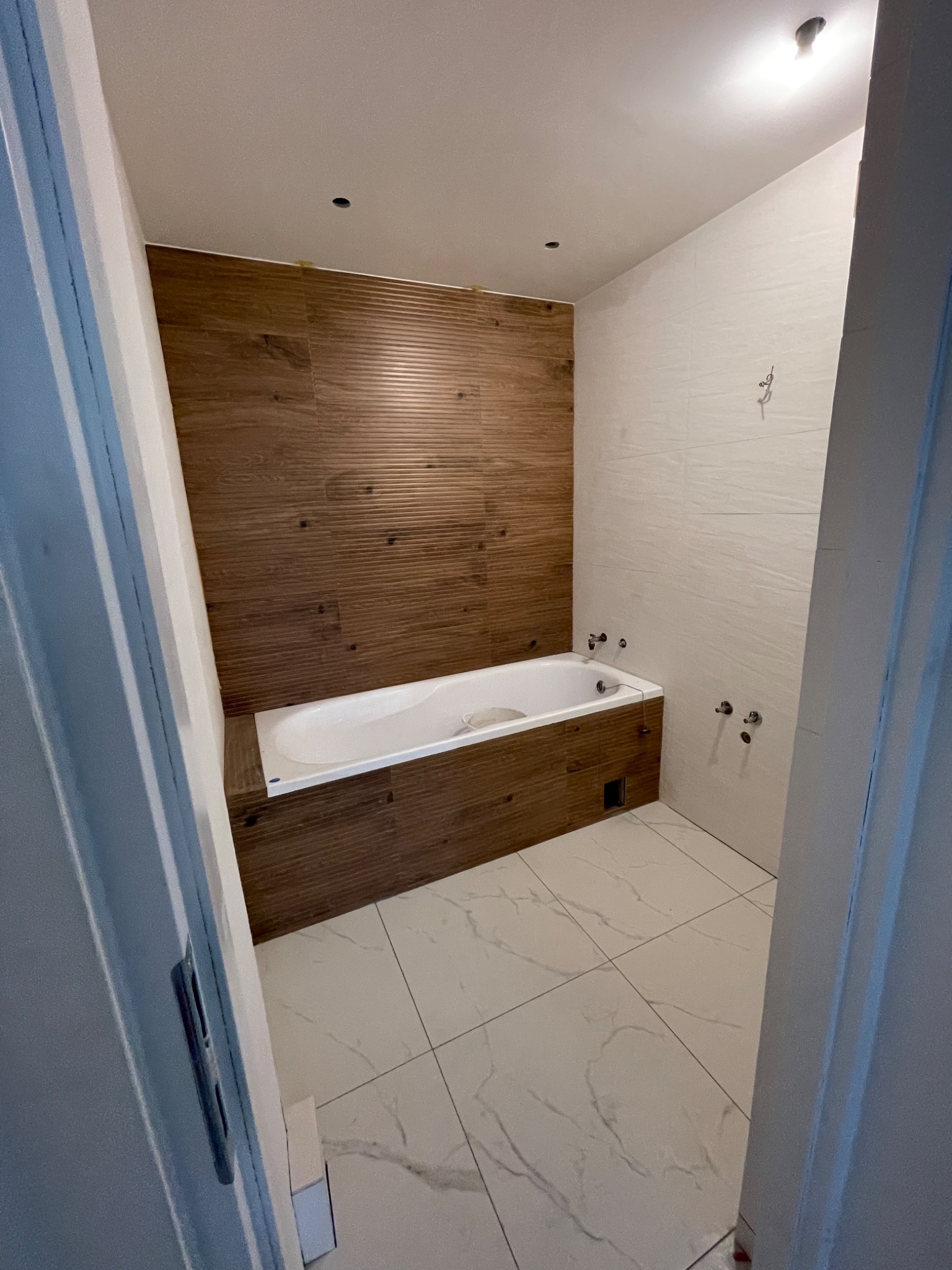 Wnętrze łazienki w trakcie remontu: biała wanna obudowana drewnopodobnymi panelami, białe płytki podłogowe z szarym żyłkowaniem, ściana wyłożona białymi płytkami, widoczne niedokończone instalacje...