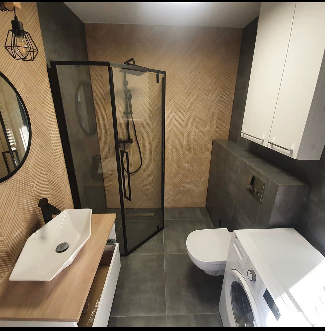 Nowoczesna łazienka z kabiną prysznicową typu walk-in z czarnymi profilami, umywalką nablatową na drewnianej szafce, toaletą i pralką, wykończona płytkami w geometryczny wzór i szarościach.