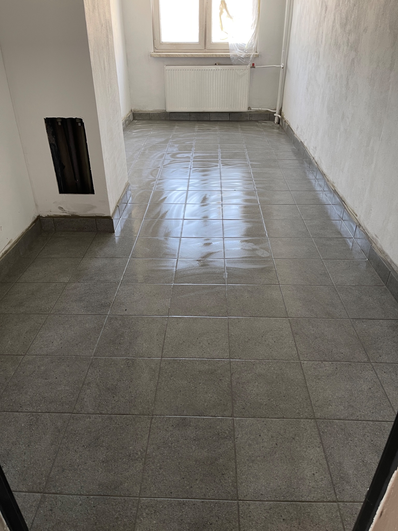 Świeżo ułożona, mokra, szara terakota podłogowa w pustym pomieszczeniu z grzejnikiem pod oknem, widoczny fragment niezabezpieczonej ściany z otworem instalacyjnym.