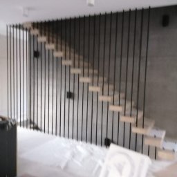 MARCIN WYKOWSKI - Nowoczesne schody z drewnianymi stopniami i czarną, pionową balustradą na tle szarej ściany, widoczne ślady prac wykończeniowych.