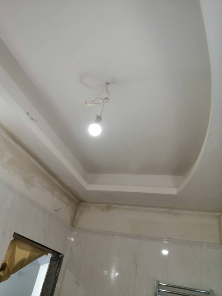 Niedokończony biały sufit z dekoracyjnym obniżeniem i wiszącą żarówką w remontowanej łazience z marmurowymi płytkami na ścianach.