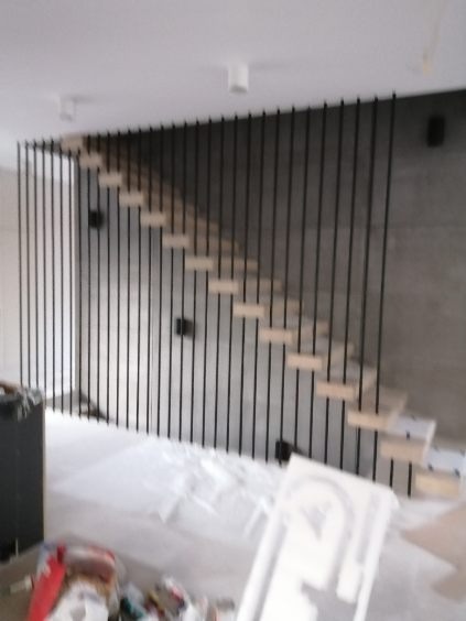 Nowoczesne schody z drewnianymi stopniami i czarną, pionową balustradą na tle szarej ściany, widoczne ślady prac wykończeniowych.