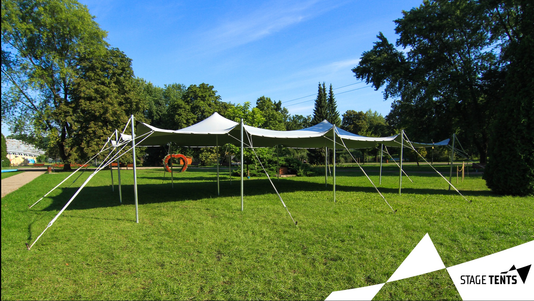 Rozłożony biały namiot stretch na trawniku, z dekoracją w kształcie serca z kwiatów, w tle drzewa i błękitne niebo.
