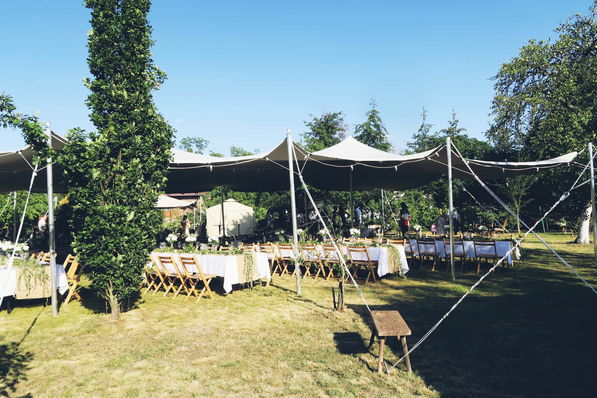 Rozległy namiot plenerowy z drewnianymi krzesłami i stołami nakrytymi białymi obrusami, udekorowanymi kwiatami i zielenią, ustawiony na trawniku w słoneczny dzień.