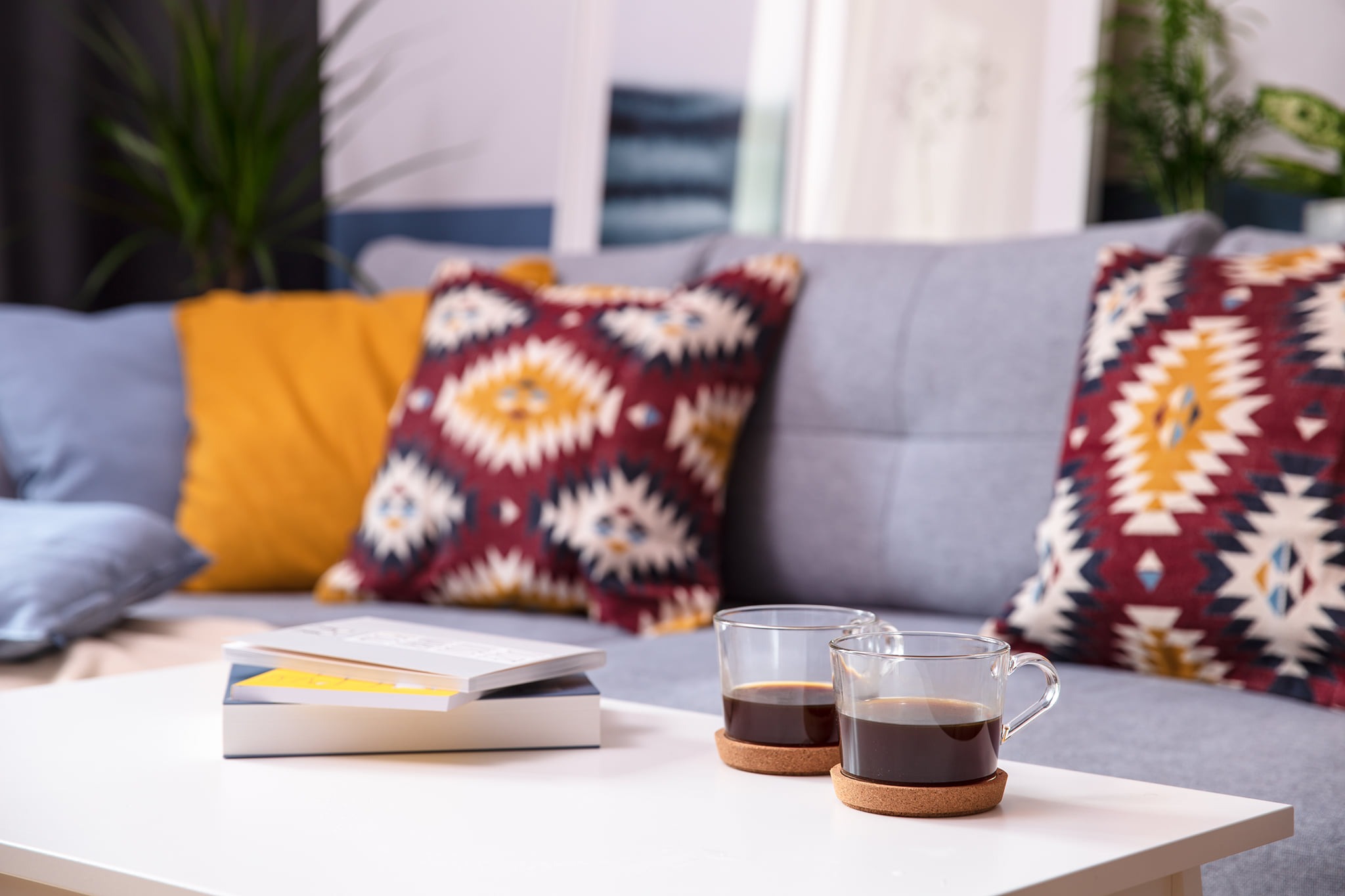 Dwie szklanki z kawą na korkowych podkładkach stoją na białym stoliku kawowym, obok leżą książki, a w tle widoczna szara sofa z kolorowymi poduszkami w stylu boho.