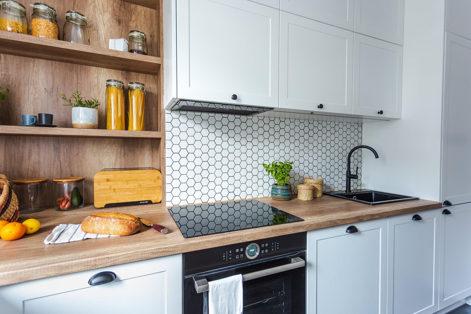 Jasna kuchnia z drewnianym blatem, białymi szafkami, heksagonalnymi płytkami na ścianie, czarnym zlewem i płytą indukcyjną, chleb na desce do krojenia.