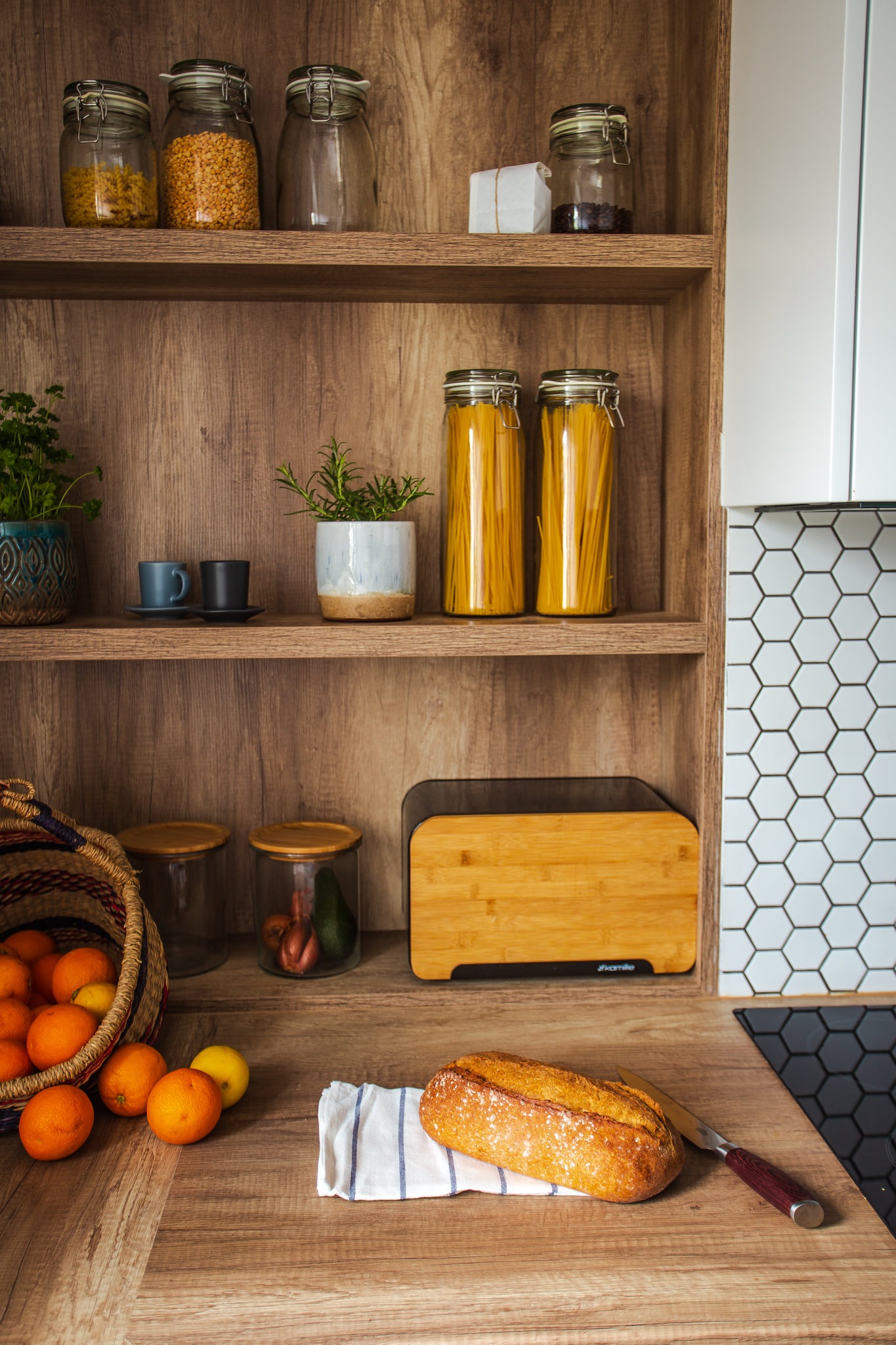 Półki w kuchni z drewnianymi elementami, na których znajdują się słoiki z makaronem, kaszą i przyprawami, obok stoi koszyk z pomarańczami i cytrynami, a na blacie leży świeży chleb na ściereczce.