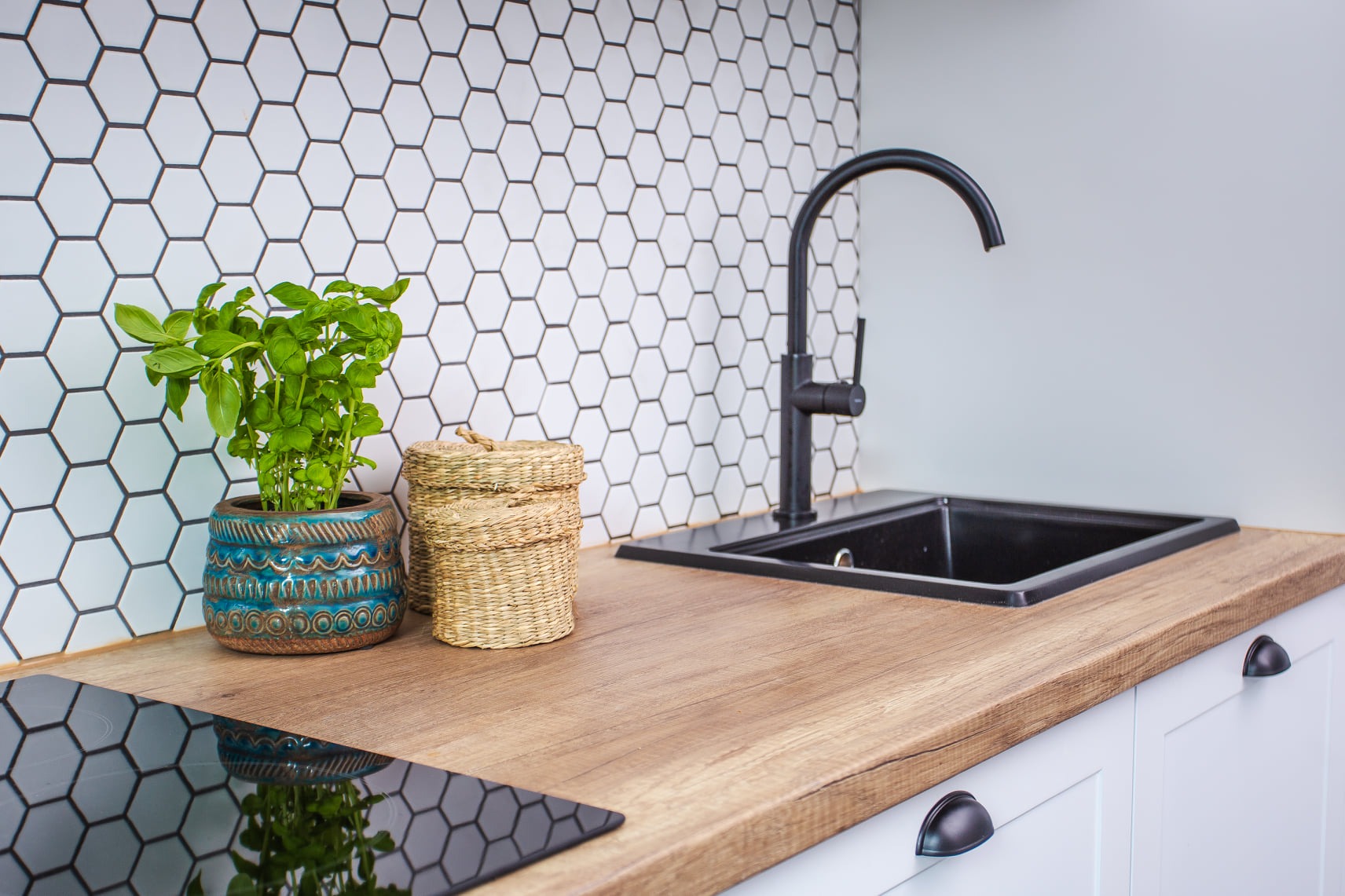 Fragment blatu kuchennego z drewnianym wykończeniem, czarny zlew i bateria, ceramiczna doniczka z bazylią, pleciony koszyk i białe heksagonalne płytki na ścianie.