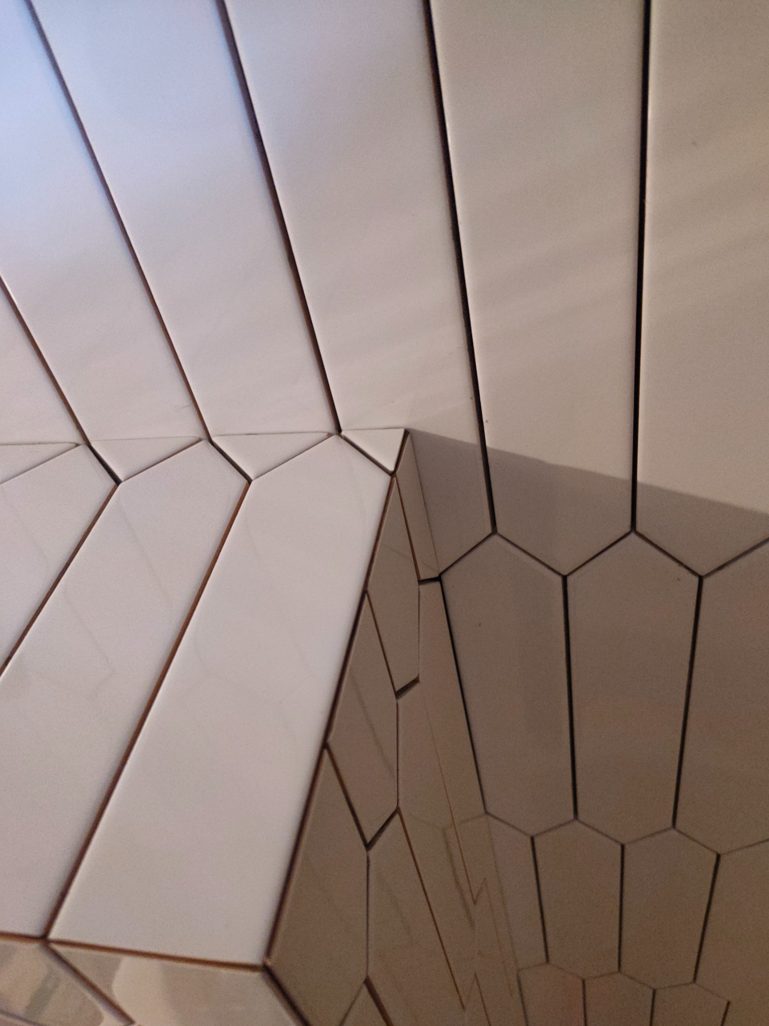 Nietypowe ułożenie białych, podłużnych płytek ceramicznych na ścianie i załamaniu rogu, tworzące geometryczny wzór z ciemnymi fugami.