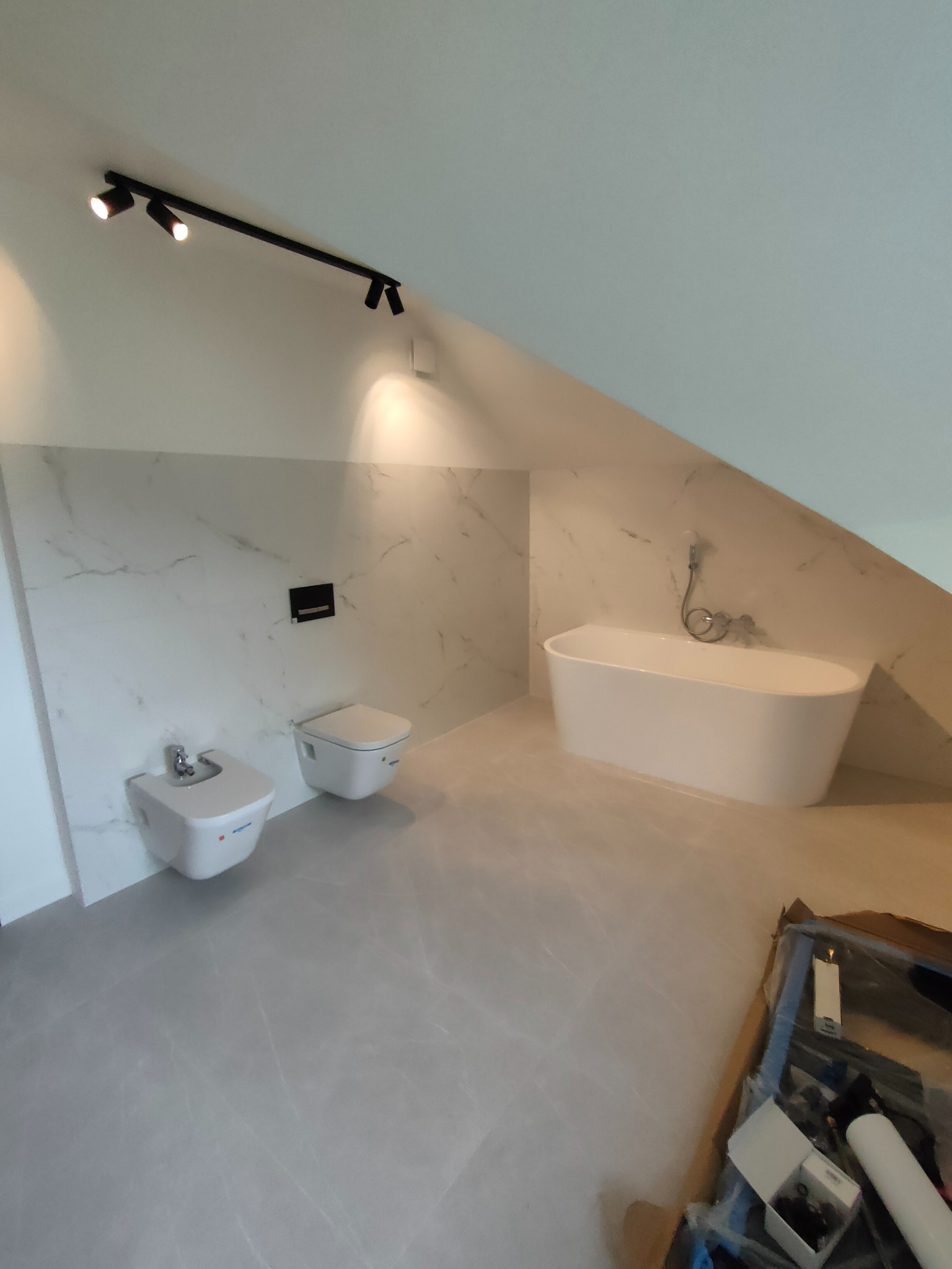 Wykończona łazienka na poddaszu w Warszawie z wanną wolnostojącą, bidetem i toaletą podwieszaną, z marmurowymi płytkami na ścianie i szarą podłogą, oświetlona czarną listwą z reflektorami.