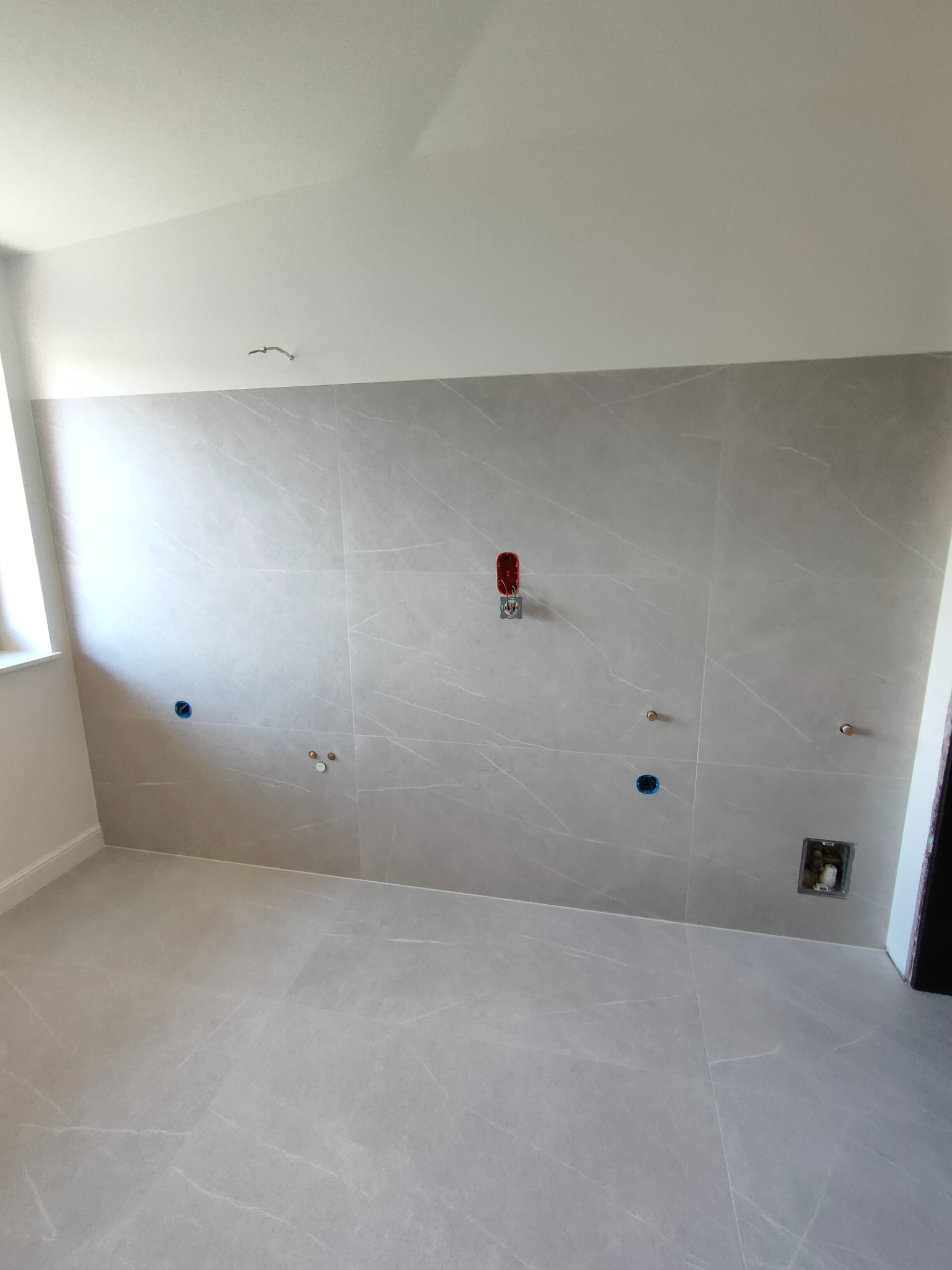 Wnętrze łazienki w trakcie prac wykończeniowych; szare płytki imitujące marmur na ścianach i podłodze, widoczne przyłącza hydrauliczne i elektryczne, brak armatury.