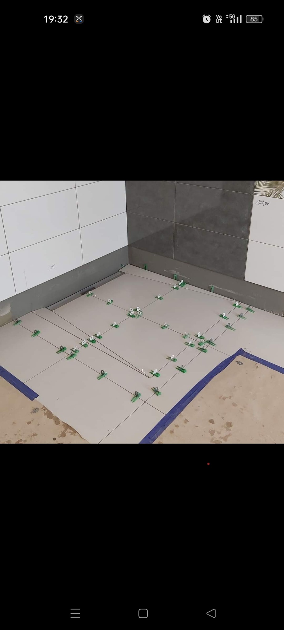 Proces układania płytek podłogowych w łazience, widoczne krzyżyki dystansowe i sznurki wyznaczające linie, dolna część ściany wykończona szarymi kaflami.