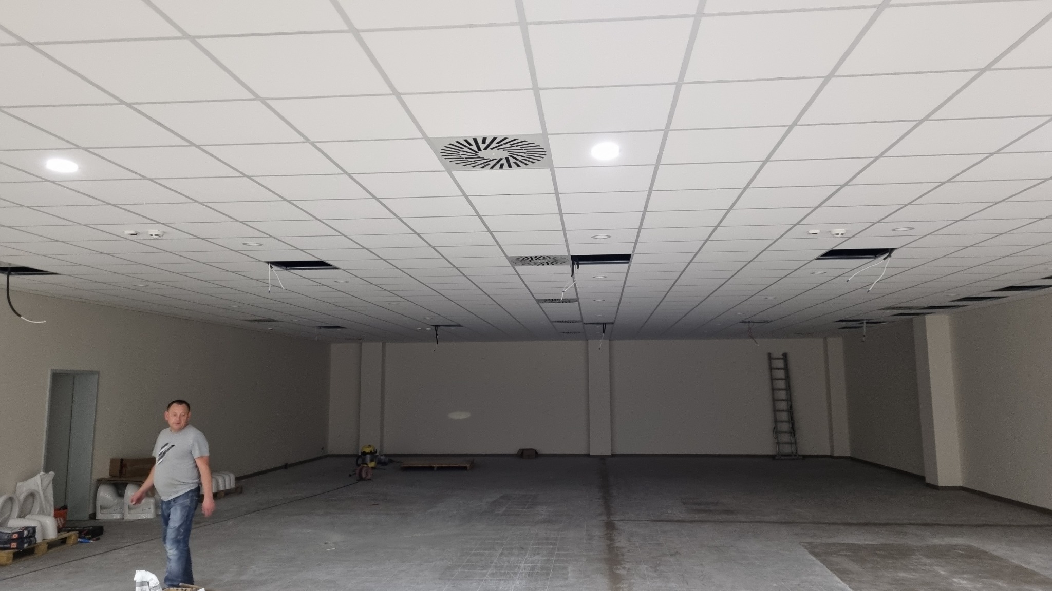 Montaż sufitu podwieszanego kasetonowego w przestrzeni biurowej, widoczne otwory na instalacje, wentylacja i oświetlenie, pracownik w tle.