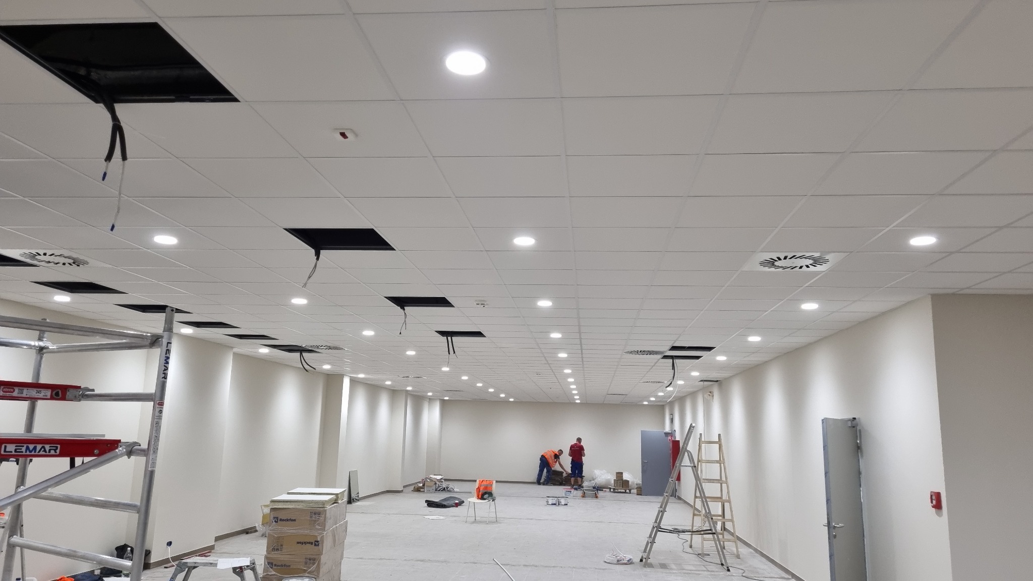 Montaż sufitu podwieszanego kasetonowego w przestronnym biurze z widocznymi otworami na instalacje, oświetleniem punktowym i pracownikami w tle.
