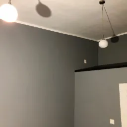 Szara ściana po malowaniu z widocznymi dwoma lampami sufitowymi i fragmentem białych drzwi z czarnymi poziomymi paskami.