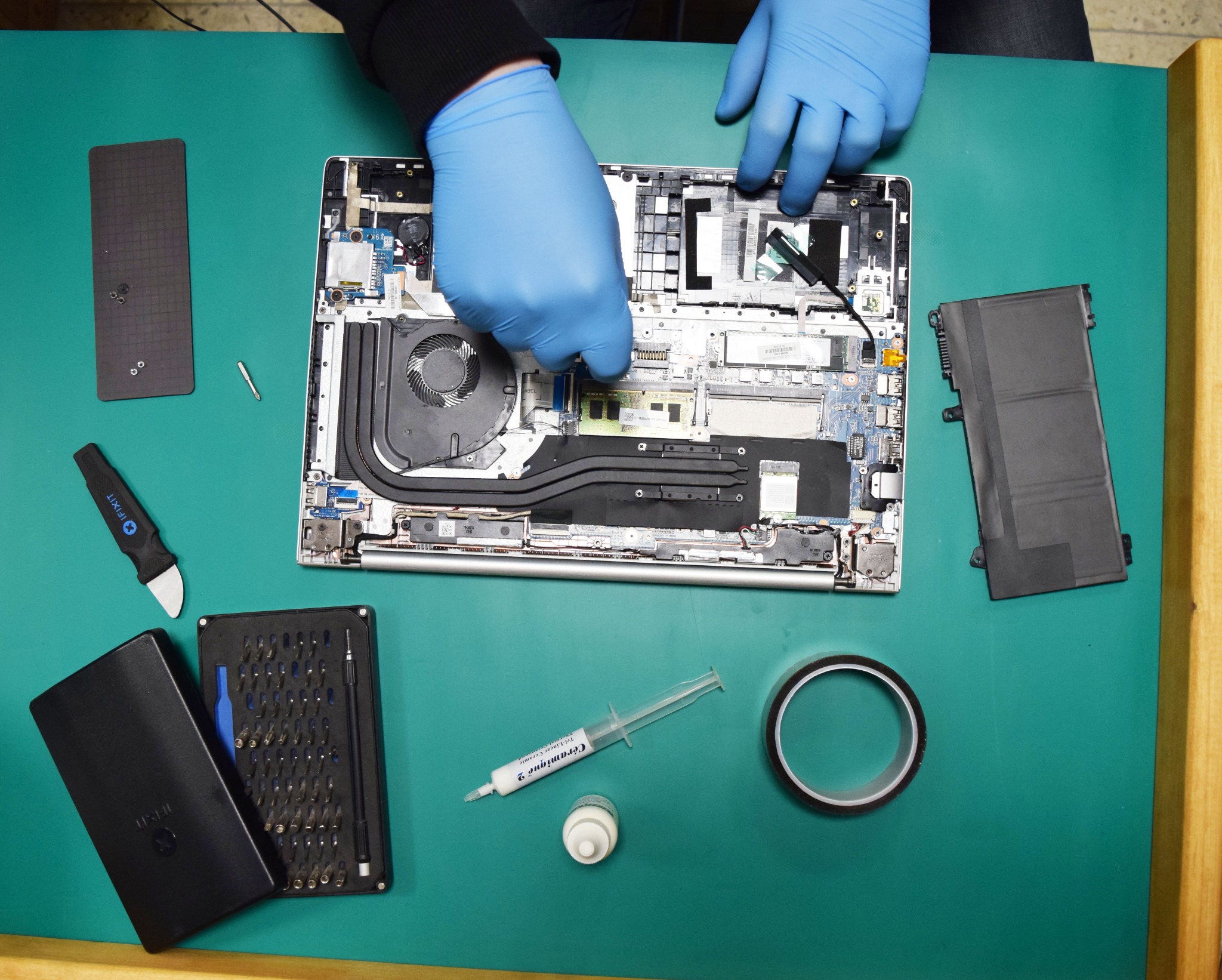 Naprawa laptopa w Gdańsku: widok z góry na otwarty laptop z widocznymi podzespołami, ręce w niebieskich rękawiczkach instalują pamięć RAM, obok narzędzia i akcesoria serwisowe.