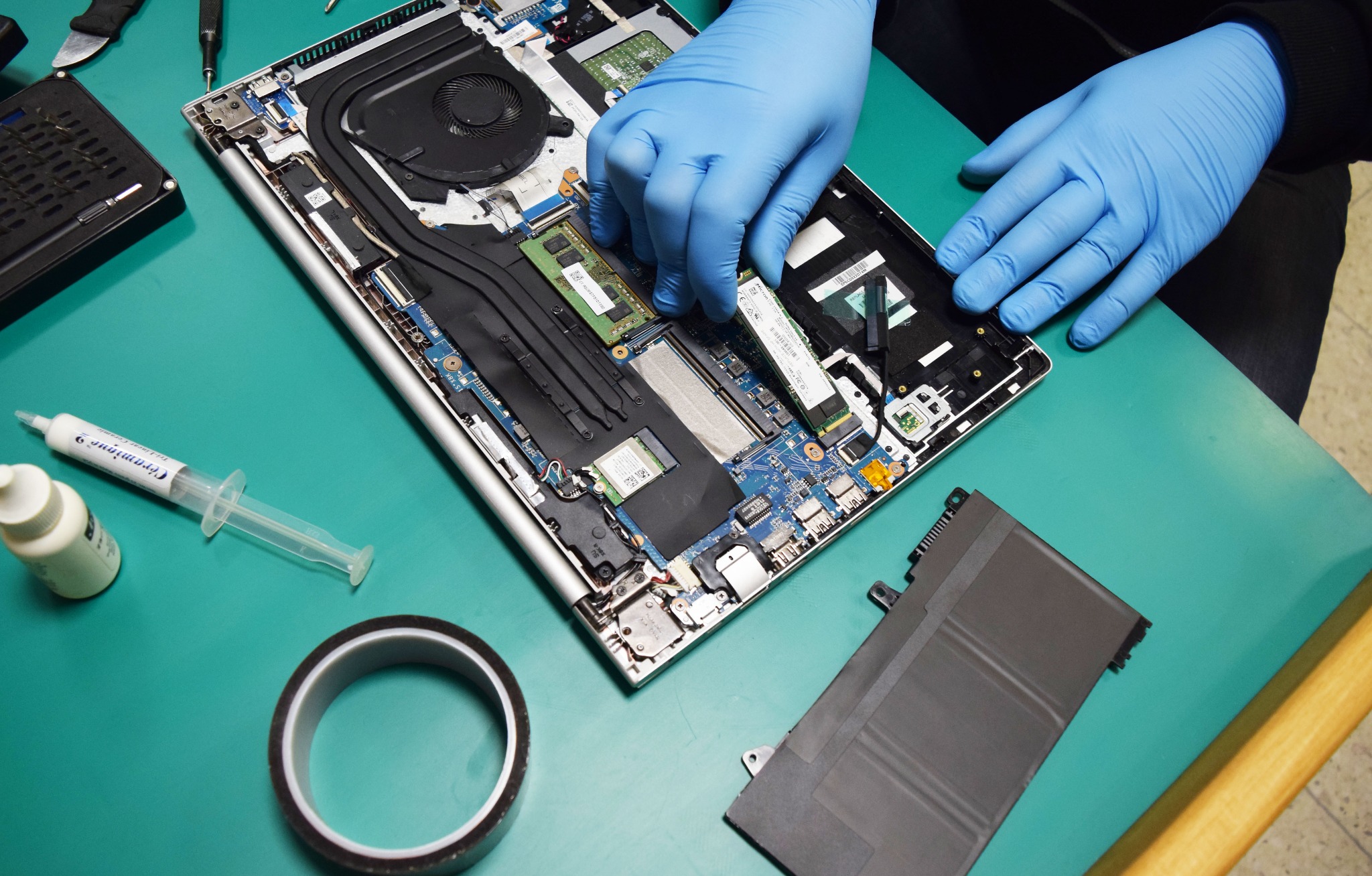 Otwarty laptop na zielonym podłożu, niebieskie rękawice lateksowe instalują moduł pamięci RAM. Widoczna bateria, pasta termoprzewodząca w strzykawce, taśma izolacyjna i narzędzia do naprawy.
