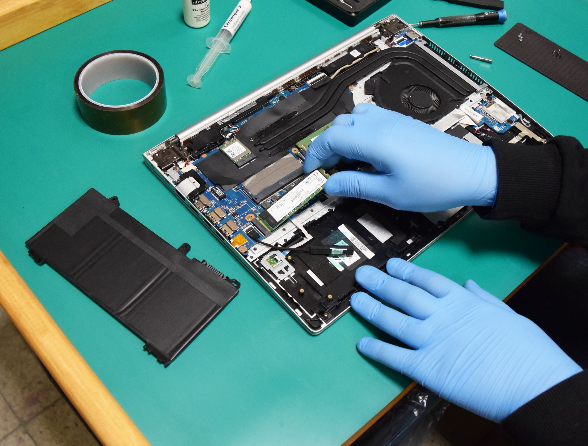 Otwarty laptop na zielonym stole, ręce w niebieskich rękawiczkach instalują moduł pamięci RAM, obok bateria, taśma izolacyjna, pasta termoprzewodząca i śrubokręty.