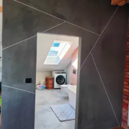 Wykończona łazienka na poddaszu z szarą ścianą dekoracyjną ozdobioną białymi liniami, widoczna pralka, wanna i okno dachowe.