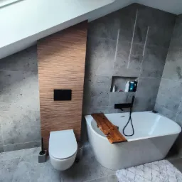 Nowoczesna łazienka na poddaszu z szarymi płytkami, wolnostojącą wanną z drewnianą deską, toaletą i drewnianą ścianką działową z czarnym panelem sterującym spłuczką.