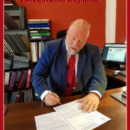 Mężczyzna w garniturze, siedzący przy biurku, podpisuje dokumenty; w tle regał z segregatorami i monitor komputera.