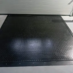 Metalowa platforma najazdowa z antypoślizgową fakturą diamentową, zamontowana przed bramą garażową z szarymi panelami, zabezpieczona słupkami z ż&oacute;łto-czarnymi pasami.