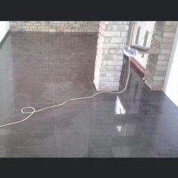 Michal Romanowicz - Błyszcząca, ciemna podłoga wyłożona płytkami, z widocznymi odbiciami światła, oraz fragment ściany z cegły i biały kabel leżący na podłodze.