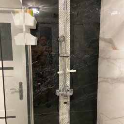 Michal Romanowicz - Nowoczesna kabina prysznicowa z czarną marmurową ścianą i srebrnym panelem prysznicowym, widoczne odbicie osoby robiącej zdjęcie w szklanej ściance.