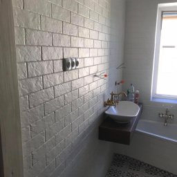 Michal Romanowicz - Biała łazienka z płytkami imitującymi cegłę, wisząca umywalka z złotą armaturą na tle wanny i okna, podłoga w czarno-białe wzory.