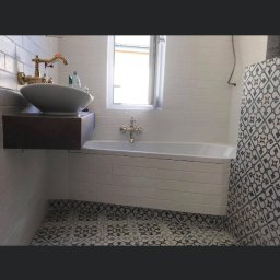 Michal Romanowicz - Nowoczesna łazienka z białą cegłą na ścianach, wolnostojącą umywalką na drewnianym blacie, baterią w kolorze złota, białą wanną i wzorzystą, czarno-białą podłogą z płytek.