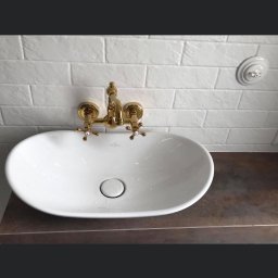 Michal Romanowicz - Biała, owalna umywalka Villeroy & Boch z baterią w kolorze złotym na blacie w odcieniach brązu, biała ściana z cegiełek w tle, widoczne krople wody.
