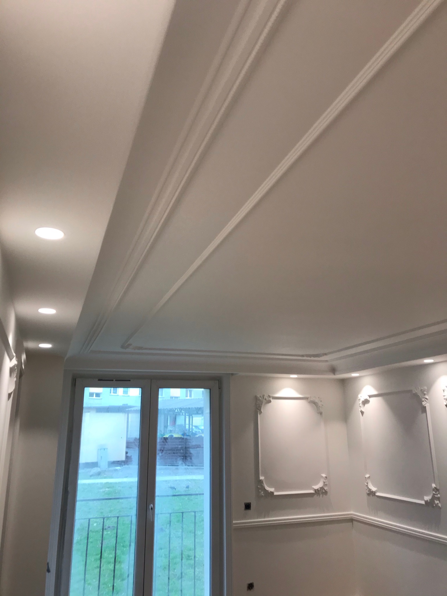 Wykończone wnętrze: biały sufit z dekoracyjnymi listwami przysufitowymi i wpuszczonymi punktowymi lampami, ściany z ozdobnymi ramami, okno z widokiem na zewnątrz.