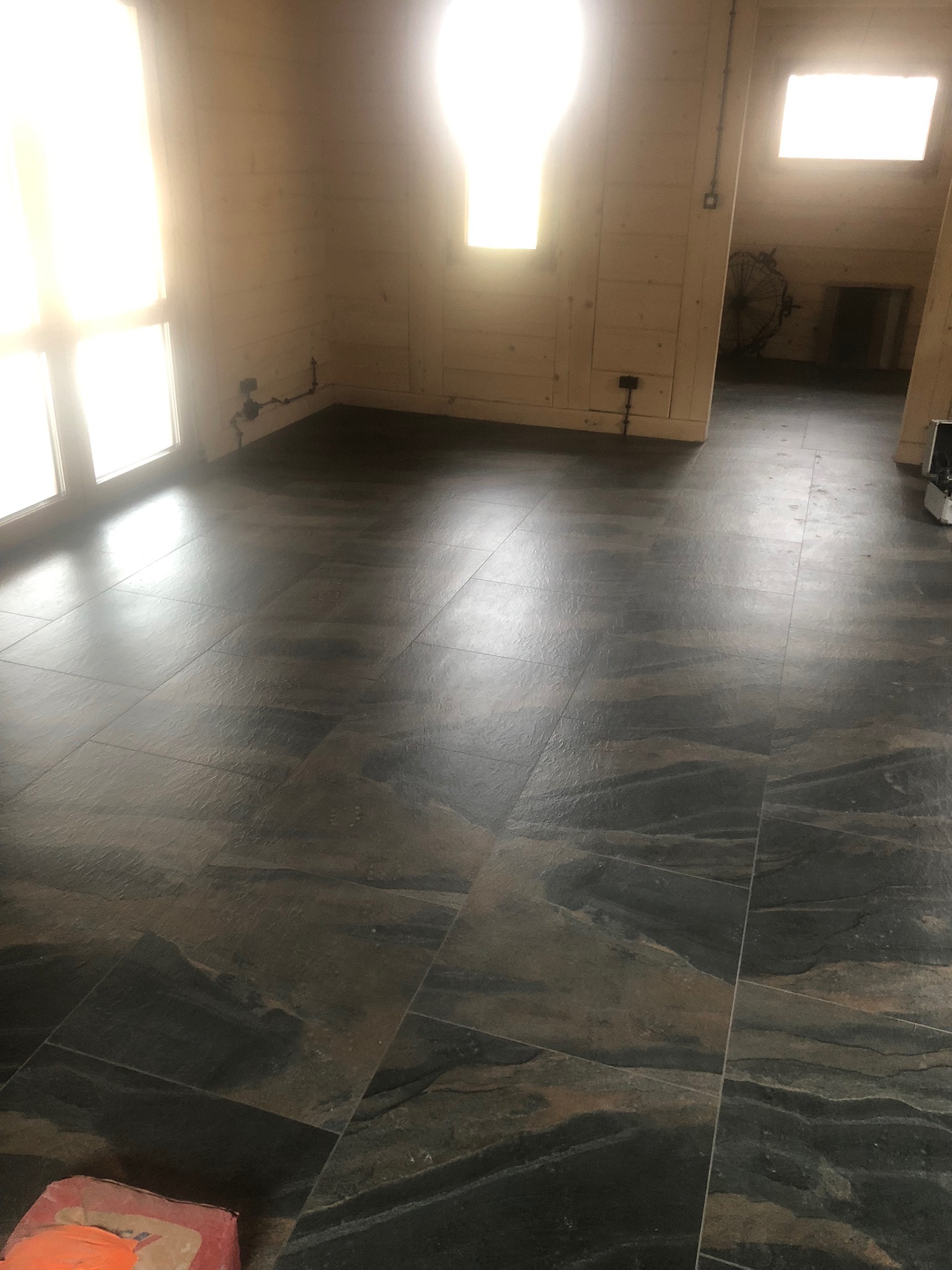 Ułożone ciemne, marmurkowe płytki podłogowe w jasnym pomieszczeniu z drewnianą boazerią, widoczne okna i fragment narzędzi.