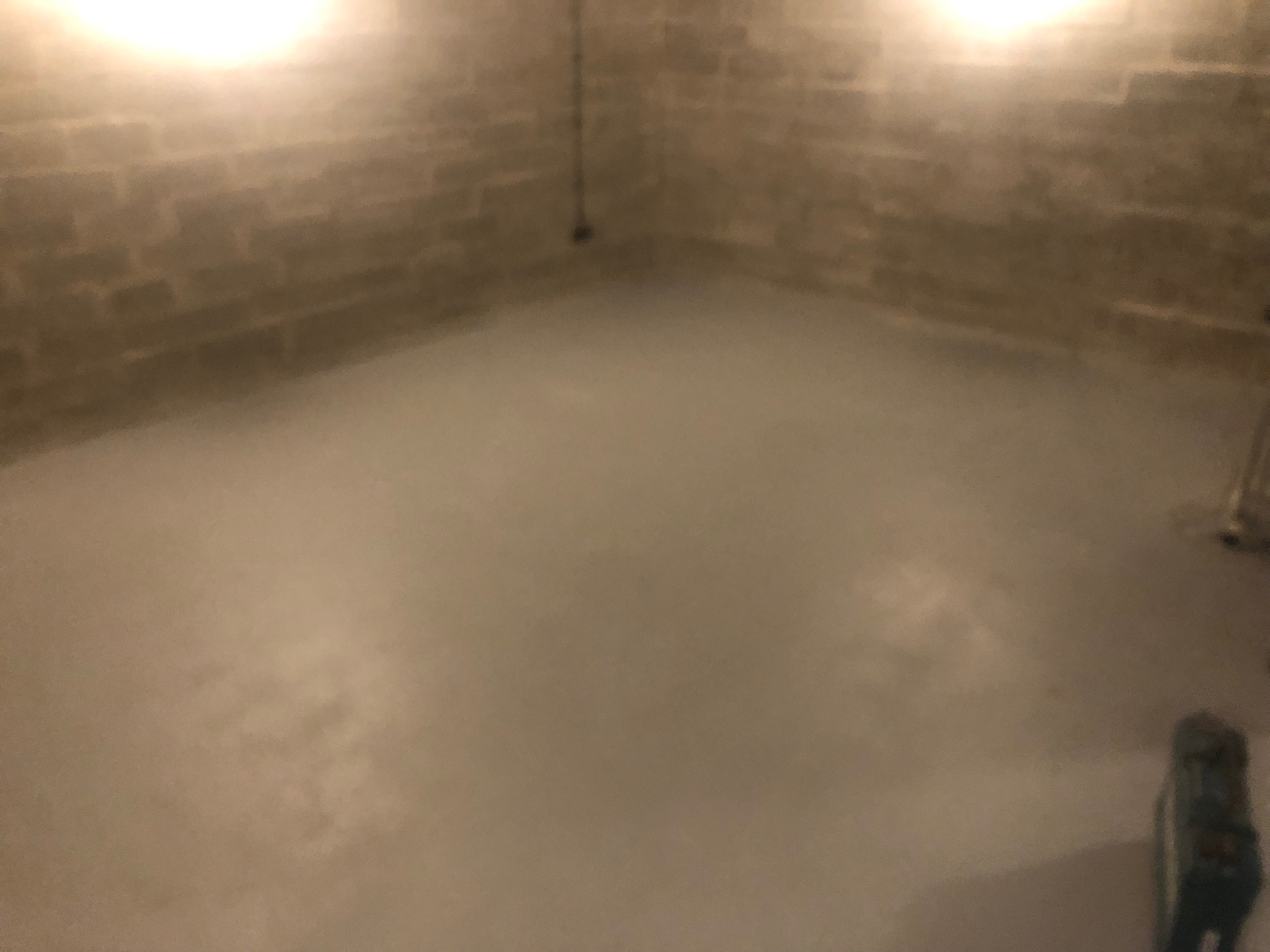 Surowe wnętrze piwnicy z betonowymi ścianami i świeżo wylaną, gładką, szarą posadzką. Widoczne ślady po szalunkach na ścianach i kabel zwisający z sufitu. Oświetlenie górne, ciepłe.