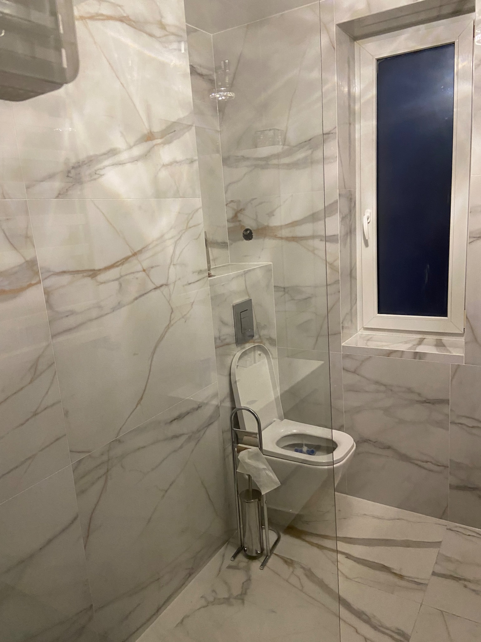 Nowoczesna łazienka wykończona białymi płytkami imitującymi marmur z delikatnymi złotymi żyłkami, kabina prysznicowa ze szkła, biała toaleta z podwieszanym systemem spłukiwania, widok na okno...