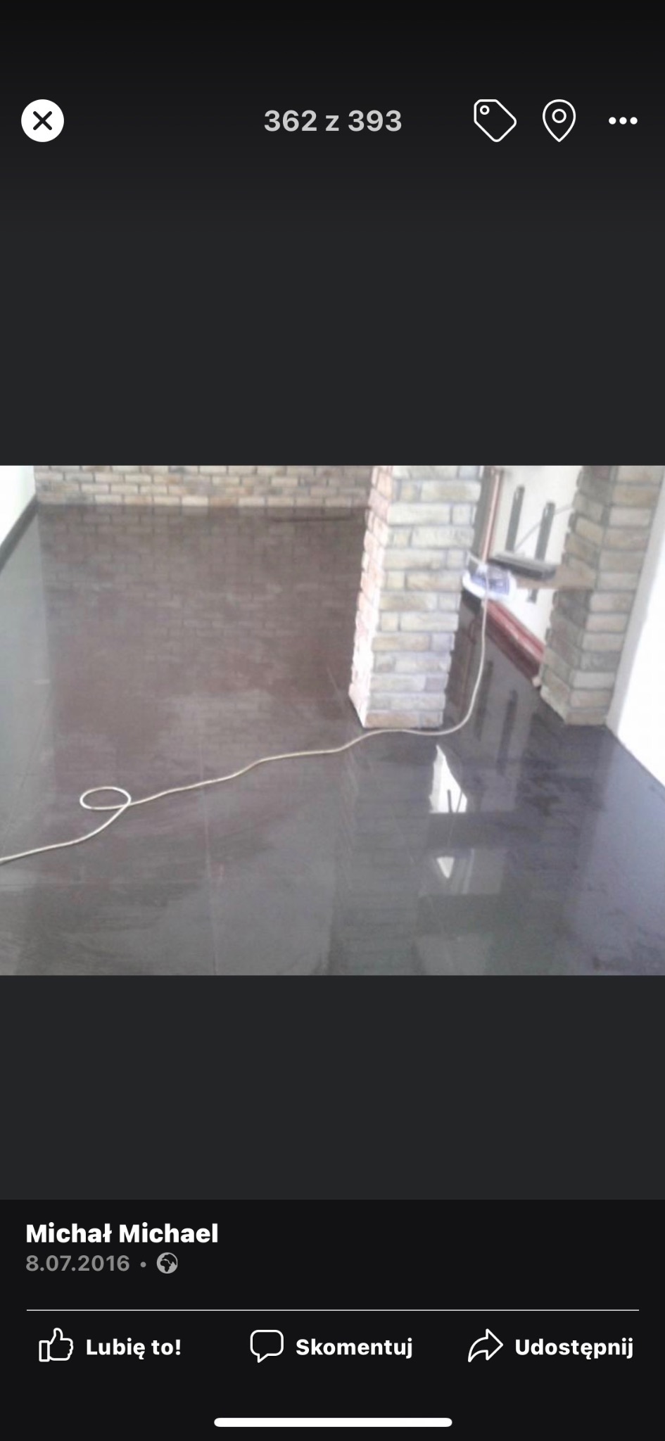 Błyszcząca, ciemna podłoga wyłożona płytkami, z widocznymi odbiciami światła, oraz fragment ściany z cegły i biały kabel leżący na podłodze.