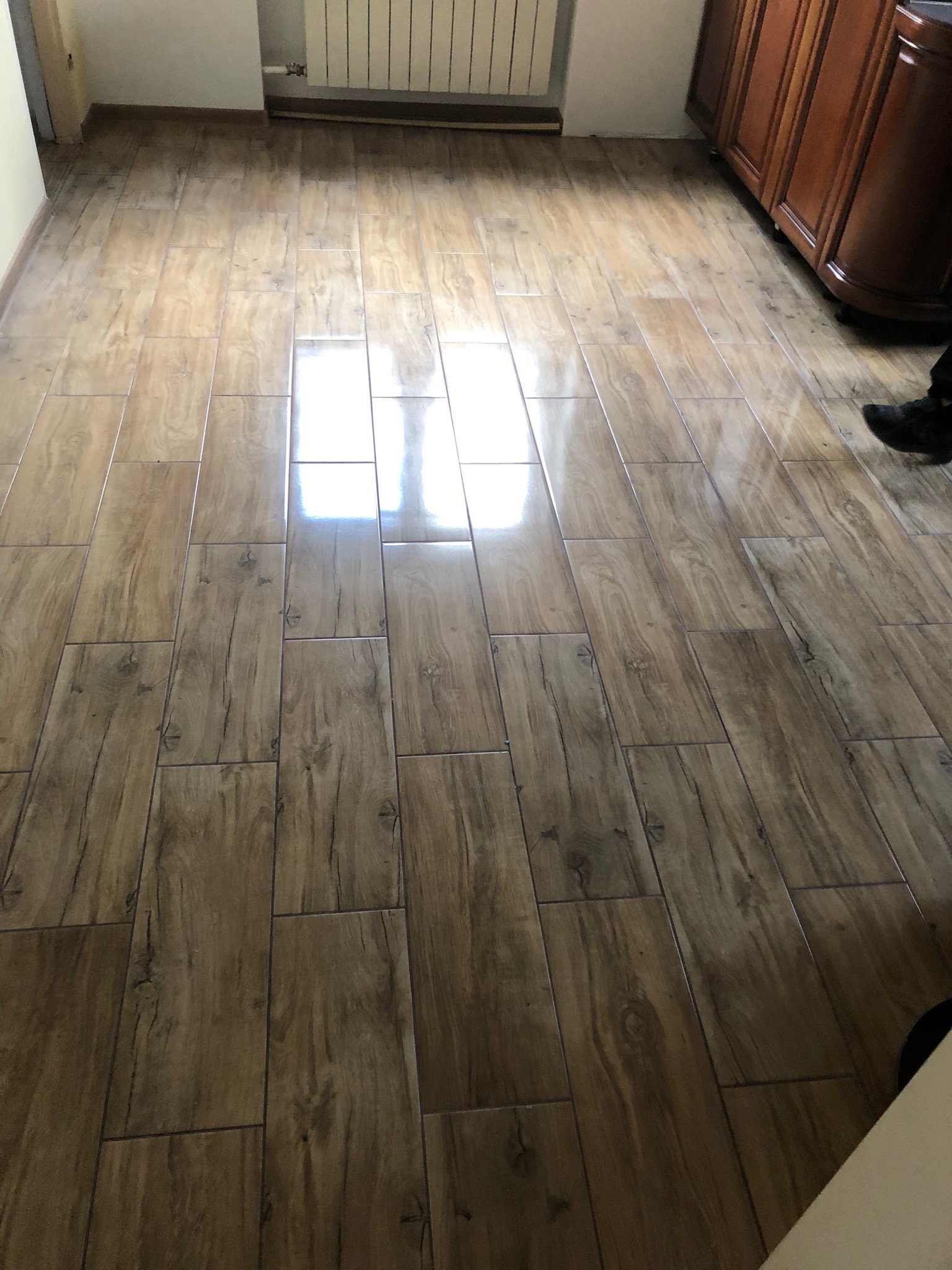 Ułożona podłoga z płytek imitujących drewno, widoczny fragment szafki i kaloryfera, błyszcząca powierzchnia odbija światło.